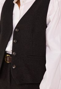 Bruine wollen gilet met vijf knopen, een enkele onderzak en een gestructureerde afwerking, gedragen over een witte blouse met opgeblazen mouwen.