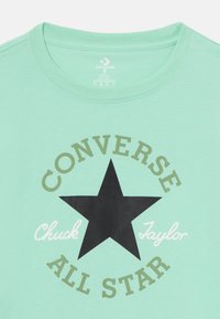 Camiseta verde menta con un gráfico de estrella negra y el texto "Converse All Star" en verde y blanco, presenta un cuello redondo y una tela suave.
