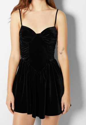 Robe en velours noir avec un corsage ajusté, décolleté en cœur et fines bretelles. Texture froncée à la taille, s'évasant en jupe.