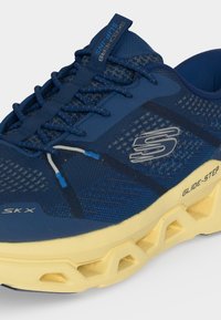 Μπλε αθλητικό παπούτσι Skechers με πλέγμα πάνω μέρος, σχέδιο με μοτίβο, μπλε κορδόνια και χοντρή κίτρινη σόλα με την επιγραφή "GLIDE-STEP."
