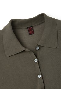 Camisa polo verde oliva con cuello acanalado, que presenta tres botones azul claro y una etiqueta de marca negra y roja en el cuello.