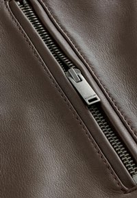 Barna bőrkabát részlete, amelyen fém cipzár található ezüst húzófüllel, cérnaszegés és textúrált felület.