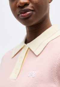 Gros plan sur une personne portant un polo en maille rose pâle avec un col et des boutons crème, arborant un petit logo blanc brodé sur la poitrine.
