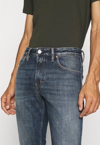 Dunkelblaue Jeans aus Denim mit leichtem Verblassen, Fünf-Taschen-Design und Kontrastnähten. Verfügt über einen Metallknopf und ein Lederpatch am Bund.