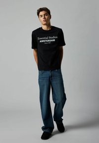 Sort bomulds T-shirt med hvid tekst: "Essential Studios AMSTERDAM Siden 1997." Sammen med løstsiddende blå jeans og sorte sko.