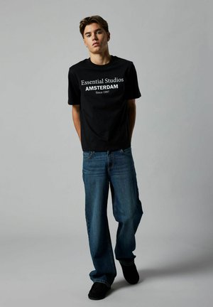 Schwarzes Baumwoll-T-Shirt mit weißem Text: "Essential Studios AMSTERDAM Seit 1997." Kombiniert mit lässigen blauen Jeans und schwarzen Schuhen.