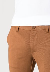 Pantalon en coton marron à coupe droite, avec une fermeture à boutons et des poches avant. Le tissu a une texture lisse.