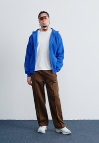 Blauwe zip-up hoodie, witte t-shirt, bruine wijde broek en witte sneakers. Accessoires omvatten oranje-getinte zonnebril.