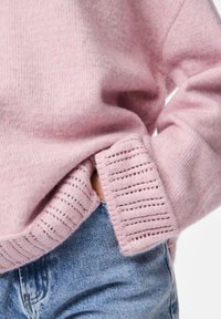 Világos rózsaszín kötött pulóver, texturált bordázott szegéllyel és mandzsetta részletekkel, világoskék farmernadrággal párosítva.