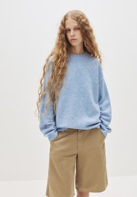 Hellblauer Pullover mit rundem Ausschnitt und gerippten Bündchen, kombiniert mit beigen Shorts. Der Stoff wirkt weich und leicht strukturiert.