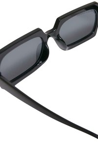 Svarta solglasögon med geometrisk design, utrustade med mörka linser och tjocka bågar. Ytan är glänsande, med en elegant och modern silhuett.