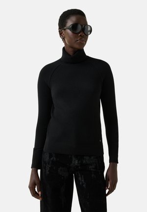Kvinde iført sort højhalset sweater, sorte mønstrede bukser og store sorte solbriller, vendt en smule mod venstre på hvid baggrund.