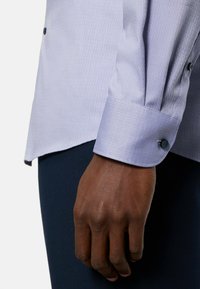 Light blue patterned dress shirt with a textured fabric, long sleeves, and a dark button on the cuff. Hand resting on a dark surface.