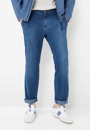 Uomo con jeans blu risvoltati, sneakers bianche con dettagli blu, giacca azzurro chiaro e camicia bianca, in piedi davanti a uno sfondo bianco.