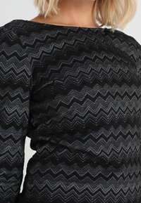 Kobieta ubrana w czarną i szaro-skalną bluzkę z długim rękawem w zygzakowy wzór, z dekoltem łódkowym i lekko marszczoną talią.