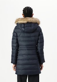 Marineblauwe gewatteerde jas met een afneembare faux fur kraag, capuchon en horizontaal gestikt ontwerp. Voorzien van lange mouwen en een lengte tot halverwege de dij.
