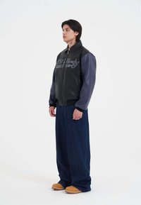 Giacca di pelle nera con maniche di pelle blu e testo ricamato, abbinata a pantaloni di jeans blu larghi e scarpe slip-on color sabbia.