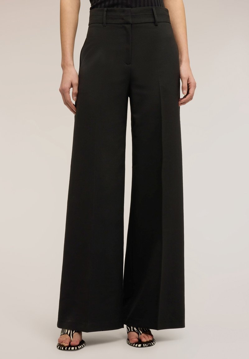 Personne portant un pantalon noir taille haute à jambes larges associé à des sandales ouvertes à rayures noires et blanches sur un fond neutre.