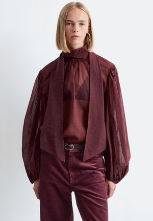 Burgunderfarbige, durchsichtige Bluse mit hohem Kragen und Puffärmeln; kombiniert mit passenden Cordhosen, die eine taillierte Taille und eine subtile Textur aufweisen.