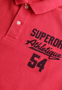 Rød polo-shirt med krave, prydet med sort broderet tekst "SUPERDRY Athletique 54" og to frontknapper. Struktureret stof.