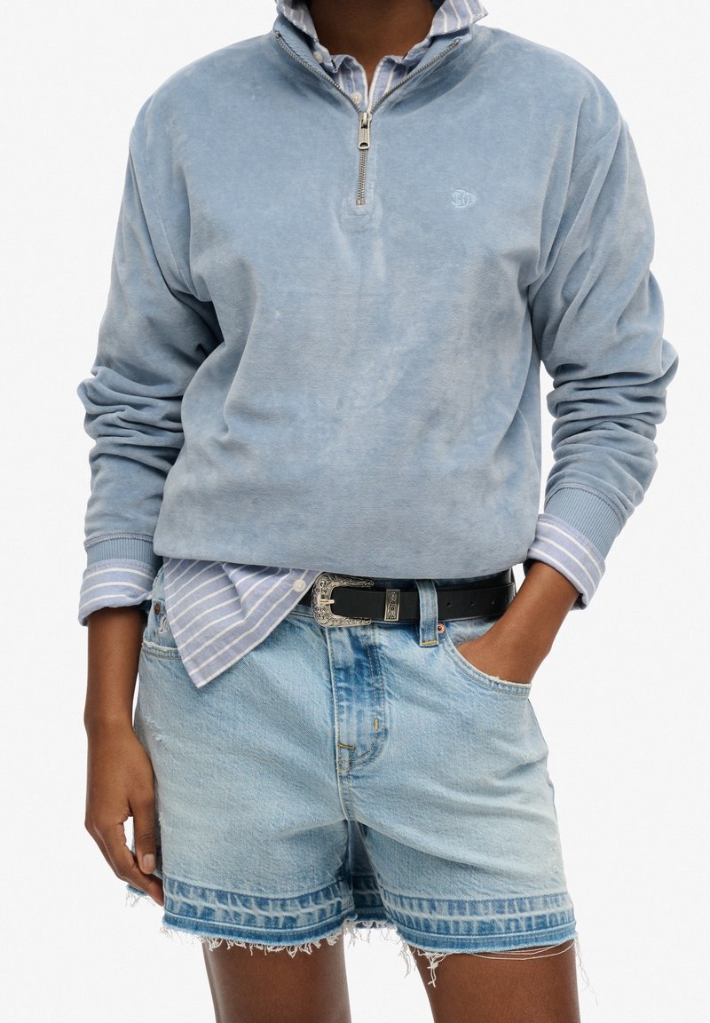 Personne portant un pull à fermeture éclair jusqu’au quart bleu clair sur une chemise rayée à boutons, associé à un short en jean clair et une ceinture noire.