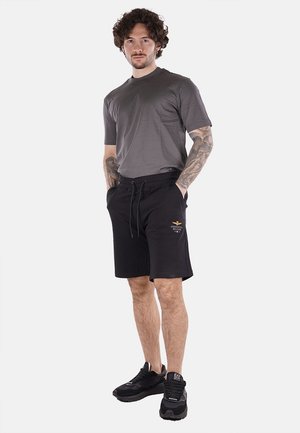 Pantaloni corti neri con cordino, caratterizzati da un piccolo logo dorato. Abbinati a una maglietta grigia aderente e a scarpe sportive nere. Il modello è in posizione frontale.