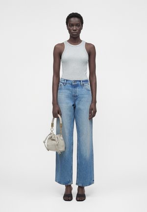 Naine seisab, kandes halli varrukateta topi, hele siniseid laia säärega teksapükse, musti avatud varbaga kingi ning hoides väikest beežika käekotti.