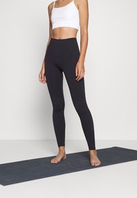 Czarne legginsy z wysokim stanem, wykonane z gładkiego materiału elastycznego, w zestawie z białą, krótką bluzką na ramiączkach, na szarej macie do jogi.