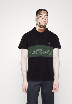 Polo negro con una franja horizontal verde que presenta "LACOSTE" en blanco. Mangas cortas y cuello clásico. Textura de tejido suave.