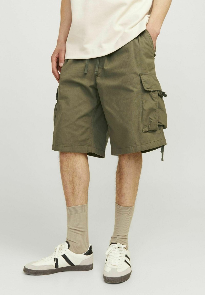 Oliwkowe szorty cargo z bocznymi kieszeniami, regulowanym sznurkiem w talii i długości do kolan, sparowane z białymi butami i beżowymi skarpetami.