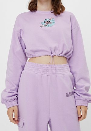 Person wearing lilac cropped sweatshirt and matching high-waist pants with a cartoon character on the chest and "Blissmind" text on pants.