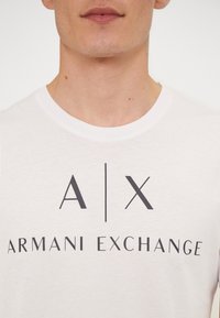 Hvit bomulls t-skjorte med rund hals, med en mørk grå "A|X ARMANI EXCHANGE" logo trykt på forsiden. Glatt tekstur.