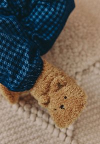 Rjavkasta copata iz flisa v obliki medveda s črnimi vezenimi detajli. Nošena z modrimi karirastimi pajkicami na teksturirani kremni preprogi.