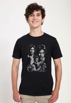 Giovane uomo con capelli ricci sorridente, indossa una T-shirt nera con più ritratti in scala di grigi di Han Solo di Star Wars.