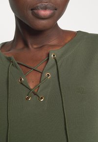 Olivově zelený top s krajkovým designem ve tvaru V, se zlatými oky a odpovídajícími krajkami. Měkká, strukturovaná látka s vyšívaným logem.