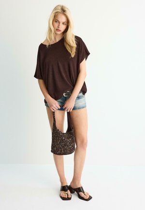 Femme blonde portant un haut marron ample, un short en denim, des sandales noires à talons, tenant un sac à main marron à motifs sur fond blanc.