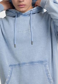 Gaiši zila kapuce, kas izgatavota no mīkstas auduma. Ietver priekšējo kabatu, regulējamus saites un izšūtu "GUESS JEANS" logotipu.