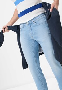 Jean bleu clair en denim, avec une coupe ajustée et des poches, associée à une veste marine tenue en main.