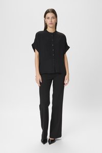 Camicia nera a maniche corte con bottoni, taglio rilassato e silhouette dritta, abbinata a pantaloni neri a gamba dritta e tacchi a punta.