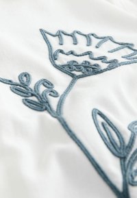 Izvezeni cvjetni uzorak u plavoj koncu na bijeloj tkanini, koji sadrži složene oblike listova i latica s teksturalnim detaljima.
