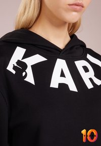 Černá mikina s kapucí s velkým bílým nápisem „KAR.“ Obsahuje malý grafický siluetu. Měkká látka s klasickým střihem a kapsou na břiše.