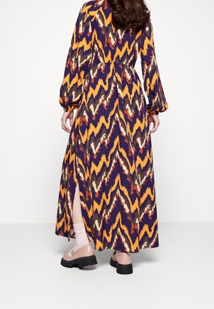 Langes Maxikleid mit Zickzackmuster in Lila, Orange und Creme. Verfügt über lange Ärmel, eine taillierte Passform und einen Seitenschlitz. Kombiniert mit klobigen Schuhen.