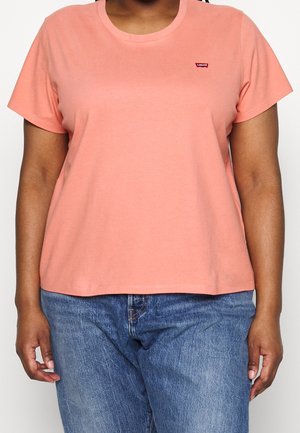 Person iført en kortærmet, lys koralfarvet Levi's T-shirt og blå denimjeans, vist fra bryst til hofter, stående mod en ensfarvet baggrund.