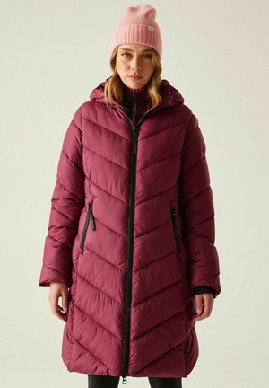 Kobieta w długim, pikowanym bordowym płaszczu z czarnymi zamkami błyskawicznymi i różową dzianą czapką, stojąca na jednolitym jasnym tle.