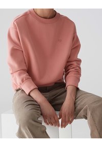 Felpa rosa chiaro con scollo rotondo e polsini a costine. Presenta un piccolo logo a forma di cuore sul petto. Tessuto morbido e vestibilità comoda.