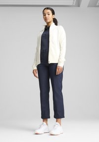 Veste blanche zippée par-dessus un haut bleu marine et un pantalon court assorti, avec une texture lisse et des chaussures de sport blanches avec des accents orange.