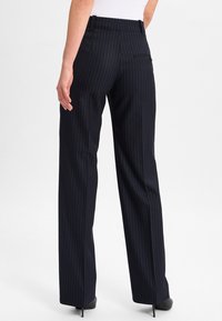 Persoon in hoge taille, donker gestreepte, wijde broek en zwarte spitse hakken, staande tegen een effen lichte achtergrond.