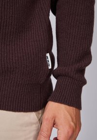 Pull en maille côtelée marron foncé avec une étiquette sur le côté, des poignets ajustés et un tissu doux et texturé.