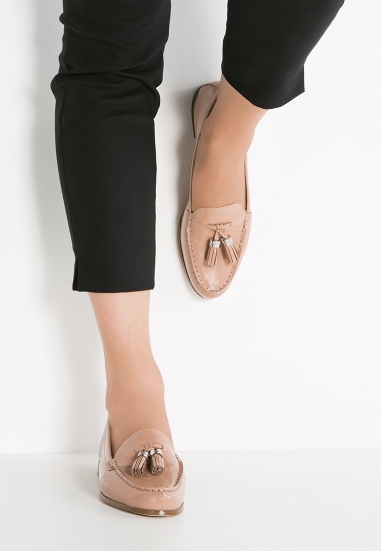 Chaussure de style mocassin beige avec une texture lisse, dotée de franges décoratives et d'accents argentés. Portée avec un pantalon noir raccourci.