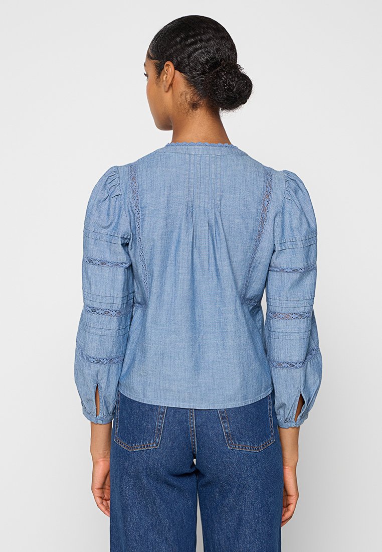 Blouse en denim bleu clair avec des manches bouffantes, des accents en dentelle et un dos boutonné. Dotée d'une coupe décontractée et de fentes sur les côtés près de l'ourlet.